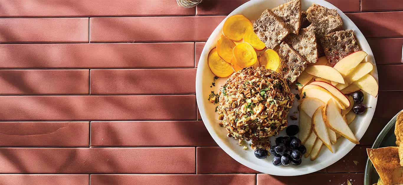 Cranberry Hummus Vegan Cheese Ball on a plate with whole grain crackers, sliced pears, and graps