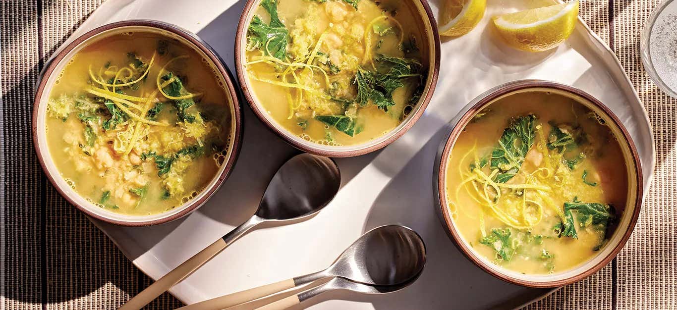 Three bowls of Lemon Hummus Soup with Kale on a tray with spoons and lemon wedges on the side