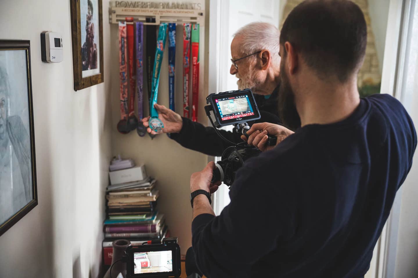 A shot from behind the scenes of the documentary More Like Paul. In the shot, 88-year-old vegan Paul Youd holds up one of his many ultramarathon medals, while a cameraman focuses on them