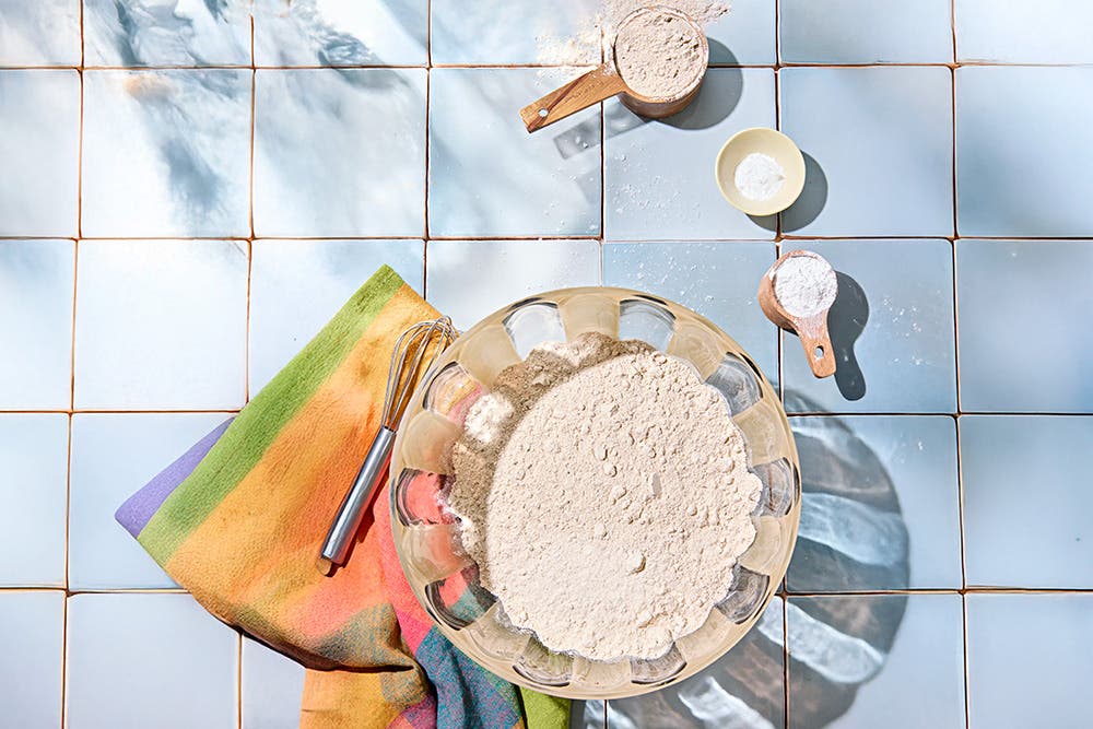 Ingredients for a healthy homemade pancake and waffle mix, with the mix in a large mixing bowl on a light blue tile surface, with a colorful kitchen towel and whisk beside it