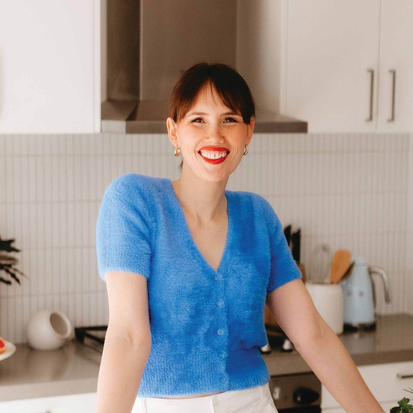 Glow Diaries founder Liz Douglas poses in her kitchen wearing a short sleeve fuzzy blue cardigan, smiling, with some vegetables on a cutting board on the counter beside her