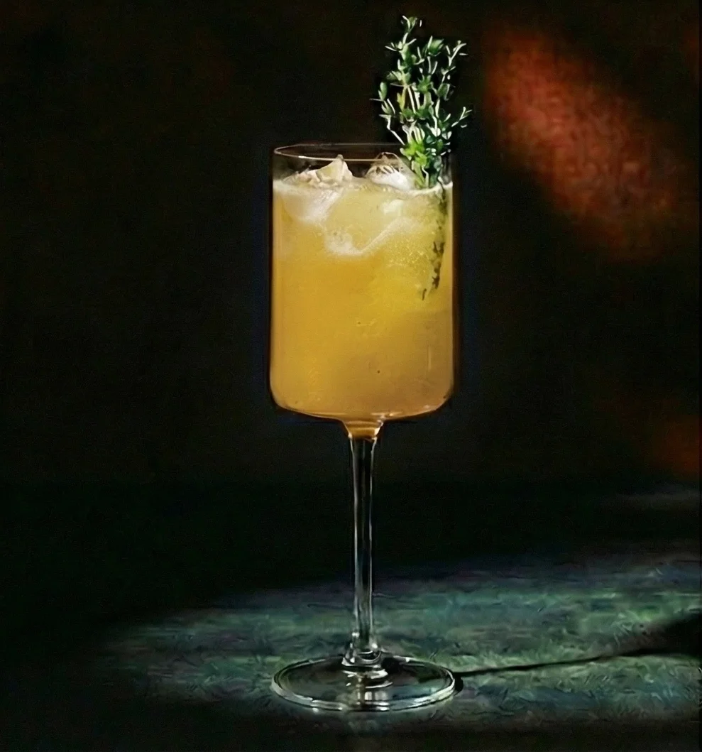 A golden-orange sparkling mocktail in a tall stemmed glass, filled with ice and topped with a sprig of fresh thyme, illuminated against a dark background.