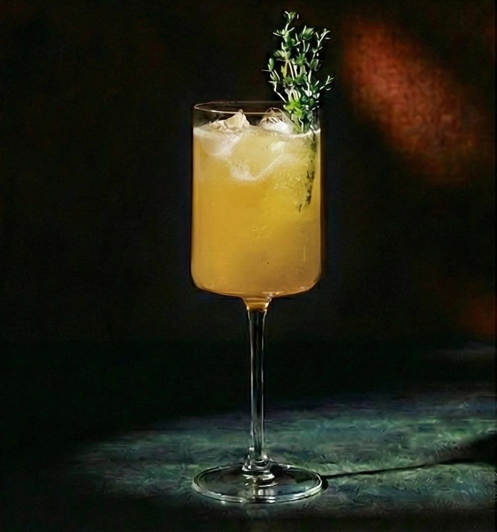 A golden-orange sparkling mocktail in a tall stemmed glass, filled with ice and topped with a sprig of fresh thyme, illuminated against a dark background.