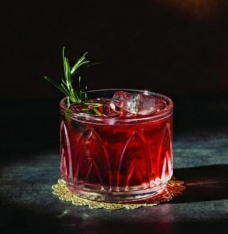 A deep red cherry spritzer served over ice in a patterned lowball glass, garnished with a fresh sprig of rosemary, set against a dark backdrop.