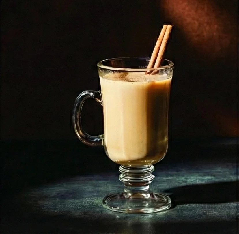 A warm, creamy chai-spiced holiday nog served in a clear glass mug with a pedestal base, garnished with two cinnamon sticks against a dark background.