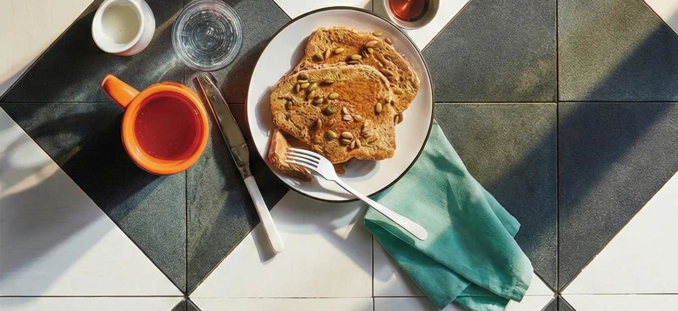 Two slices of Whole Grain French Toast on a breakfast setting with a small cup of maple syrup and a cup of tea and glass of water