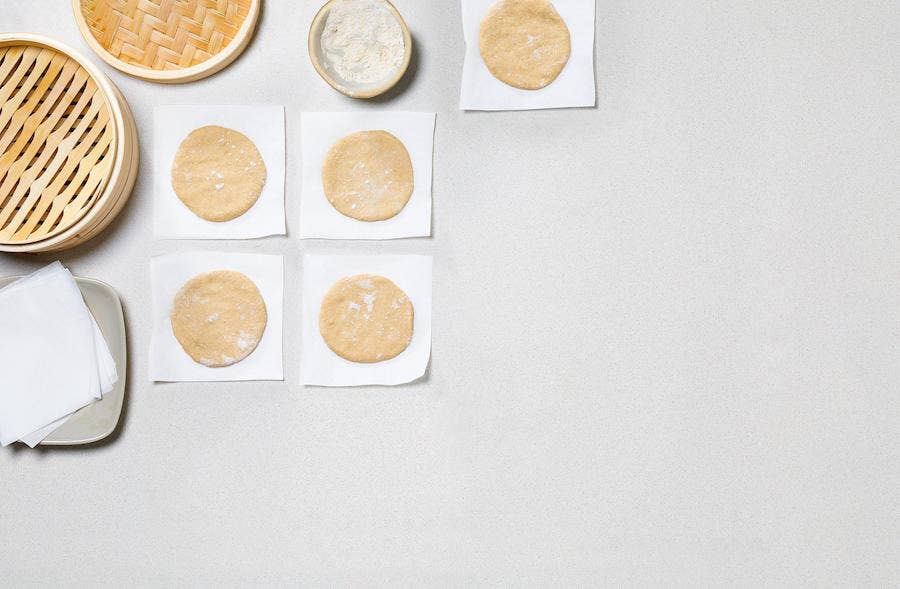whole wheat Bao bun wrappers - five circles of whole wheat dough each individually placed on a small square of parchment paper, dusted with flour, beside a bamboo steamer basket, on a white table or counter, shown from above