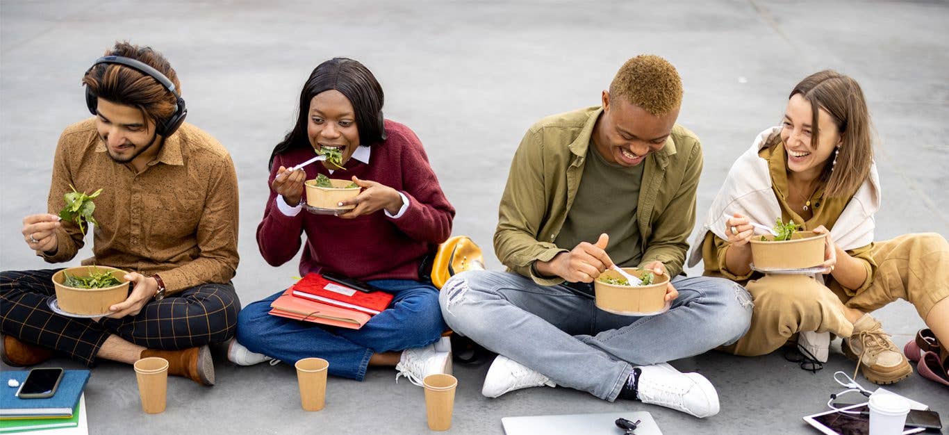 Four students of diverse backgrounds sitting and eating salad during lunch on asphalt at university campus