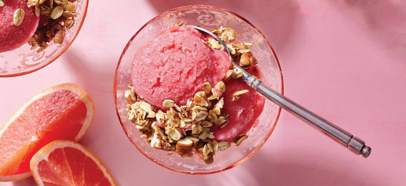 Grapefruit Nice Cream with Skillet Granola in a round glass with a metal spoon against a pink background