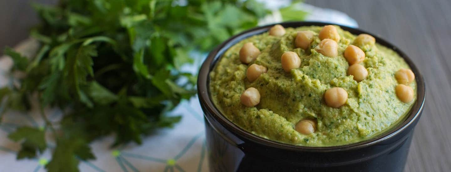 Herbed Hummus in a small black serving bowl, garnished with whole chickpeas and with a bunch of flat-leaf parsley in the background