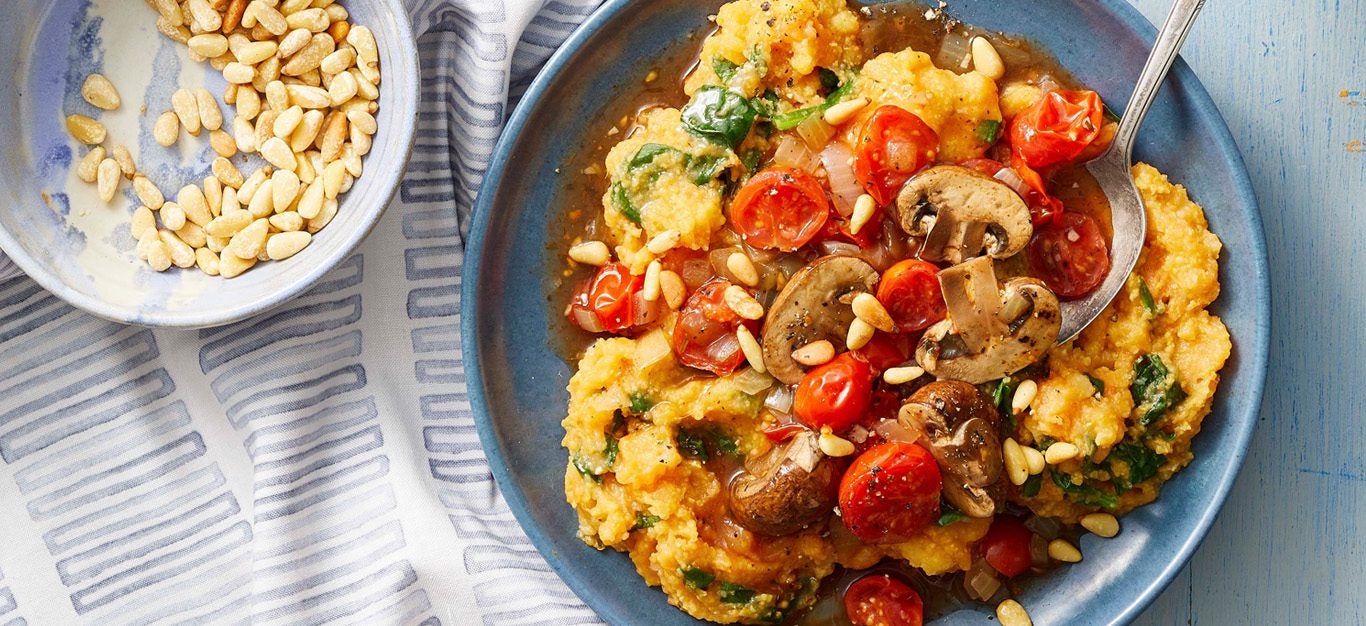 A hearty, colorful bowl of polenta and mashed sweet potato topped with blistered cherry tomatoes and juicy cremini mushrooms, with a dish of pine nuts on the side