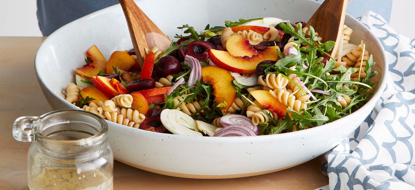 Stone Fruit Pasta Salad in white bowl with wooden salad tossers on a light wooden table