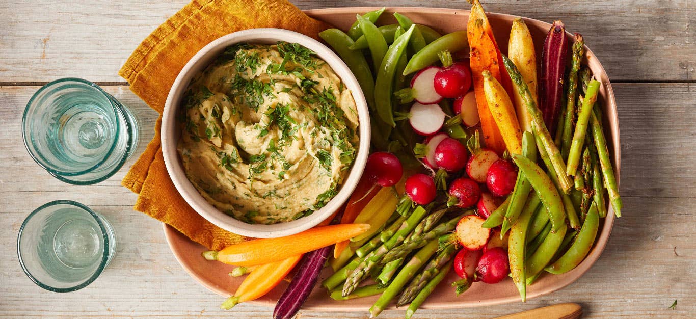 Two-Way Vegetable Platter with Herbed Hummus in a tan ceramic dish with yellow cloth napkins next to two blue water glasses