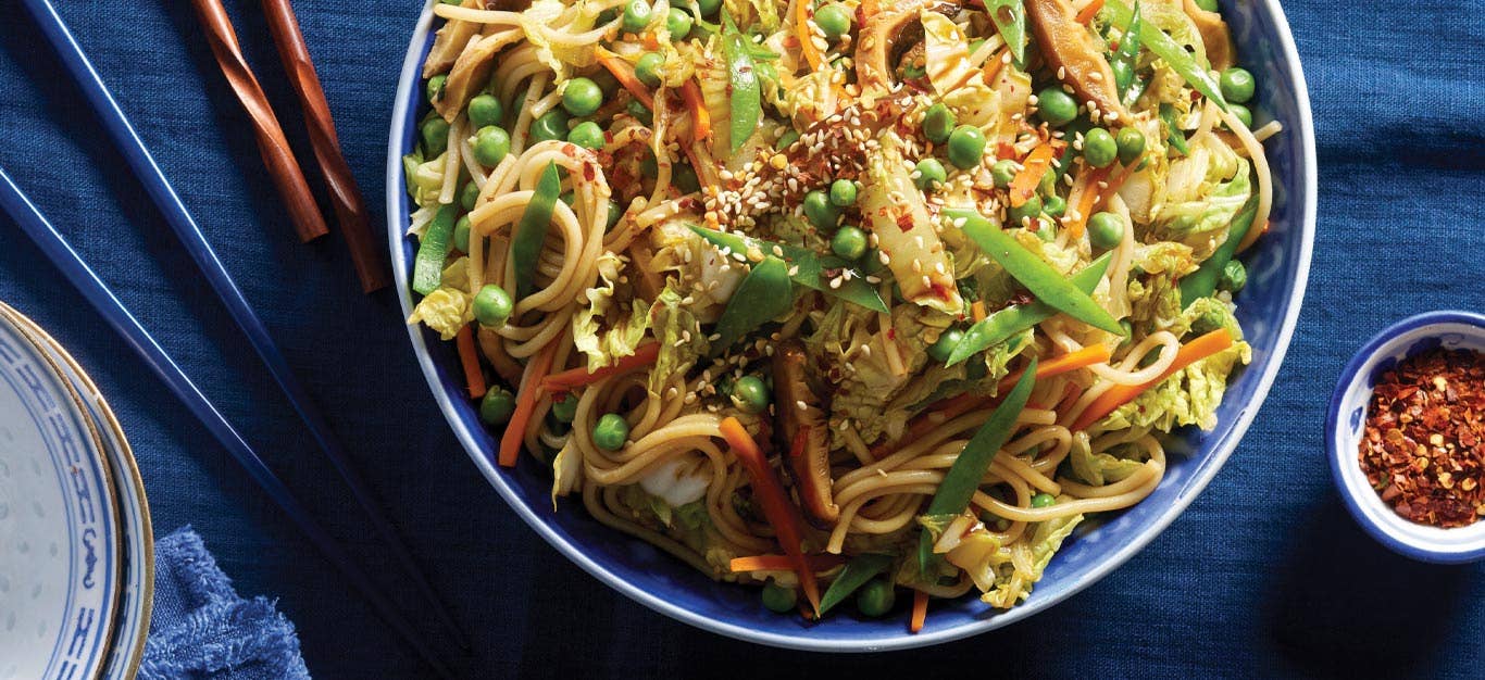 Stir-Fried Udon Noodles with Mixed Vegetables in a blue bowl with chopsticks and small bowl of red pepper flakes to the right