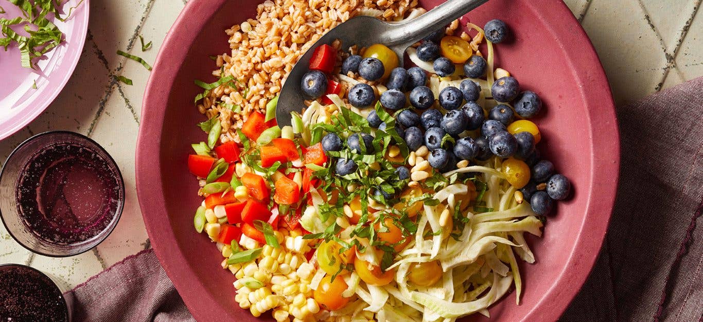 Blueberry Farro Grain Bowl - Colorful array of diced bell pepper tomatoes, fresh herbs, and corn, with farro and blueberries, in a reddish pink bowl with a glass of water beside