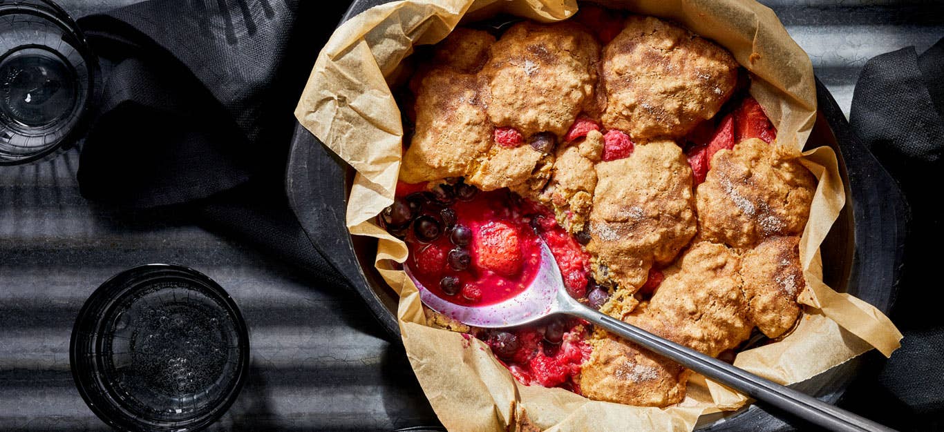 Bursting with Berries Cobbler in a cast iron skillet lined with parchment paper with a big metal spoon