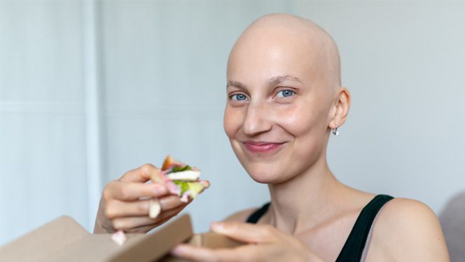 A young white woman with a bald head from undergoing cancer treatment eats a veggie flatbread