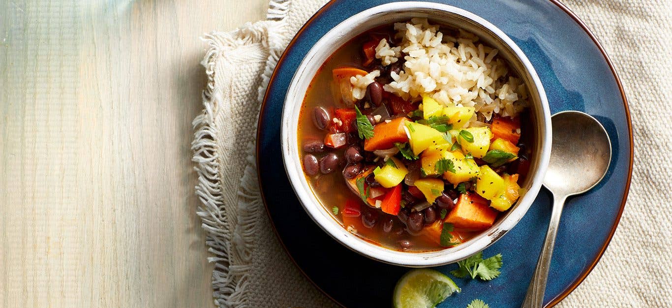 A Caribbean-inspired black bean soup, served over brown rice, topped with diced mango relish, in a white bowl atop a blue plate next to a spoon