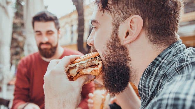 man eating processed food Young man eats a cheeseburger while another man in the background watches him
