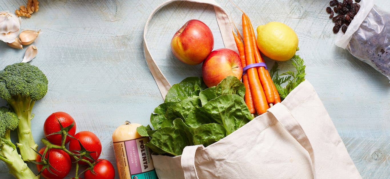 A canvas tote bag full of fresh produce lies on a white counter with more veggies surrounding it