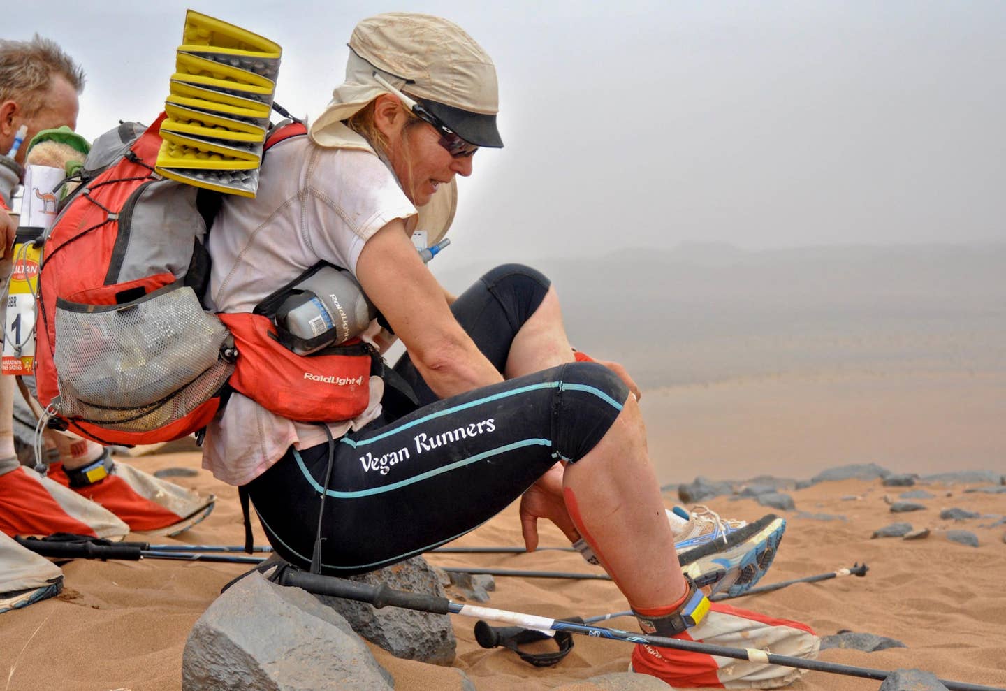 Vegan runner Fiona Oakes adjusts her shoes while wearing a backpack with a fold-up sleeping mat and a scarf around her head as she competes in an ultra marathon. On the side of her leggings is the phrase "Vegan Runners." Photo by Kirsten Kortebein