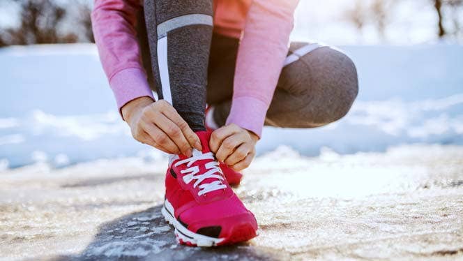 woman in a pink top and red sneakers ties her shoe on a snowy road