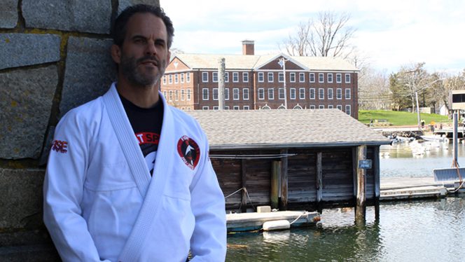 Jiujitsu martial artist Jay Oliveira stands by a stone wall in front of a small harbor