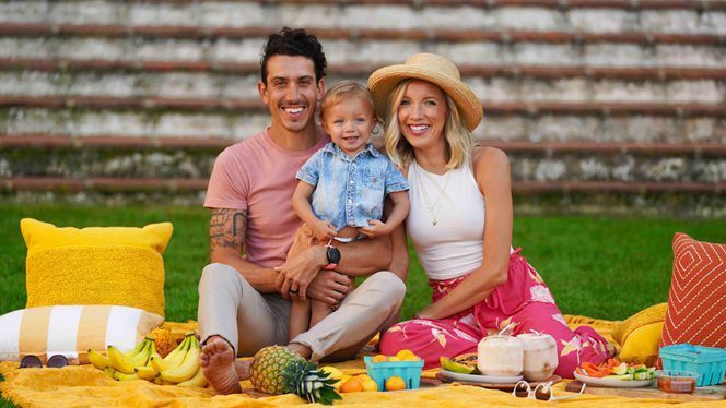 A young couple sit on a picnic blanket with their toddler between them, all smiling. Fruit including pineapple and coconut is spread out on the picnic blanket before them.