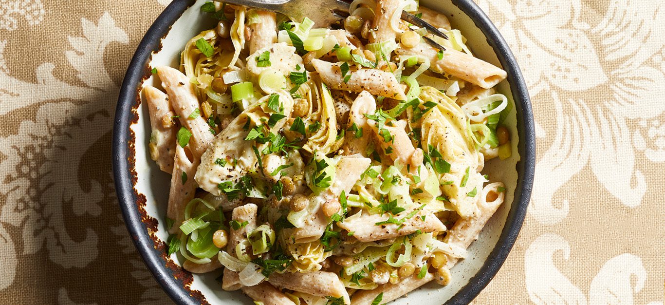 Vegan penne pasta tossed with artichokes and leeks on a white plate with black rim, sitting on a table in bright sunlight