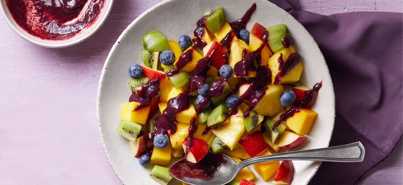 blueberry balsamic glaze sauce shown drizzled over a colorful array of fruit including kiwis and mango, on a purple table