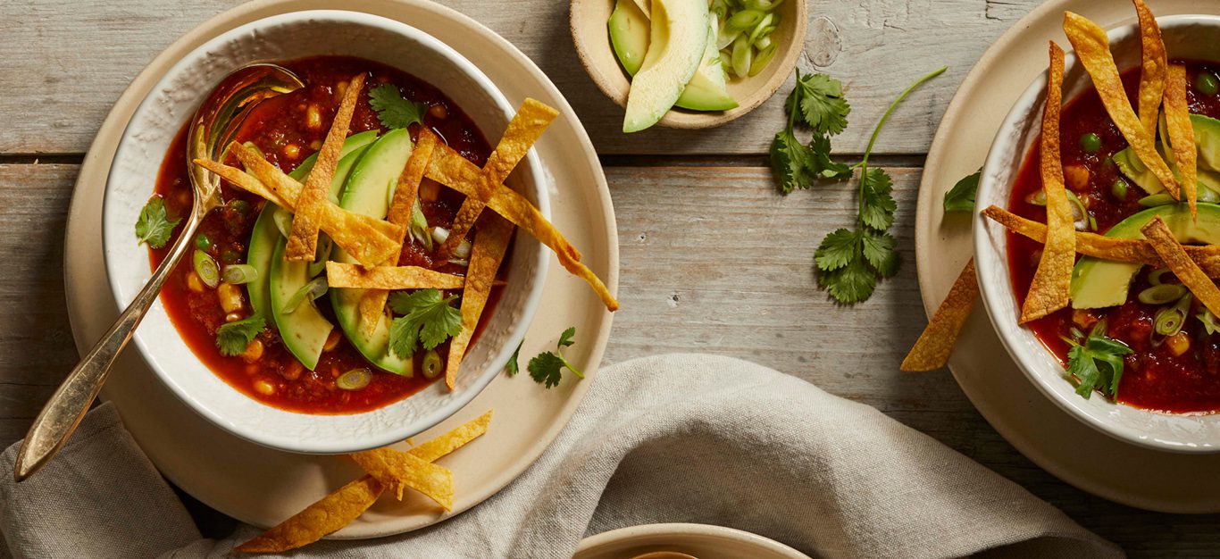 Two bowls of vegan tortilla soup topped with avocado and crunchy tortilla strips, shown on a red table with garnishes between them