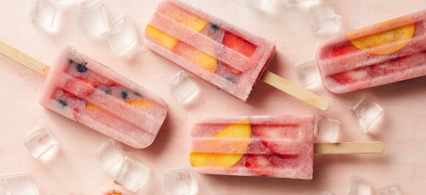 Strawberry, blueberry, and nectarine popsicles on a pink background surrounded by ice cubes
