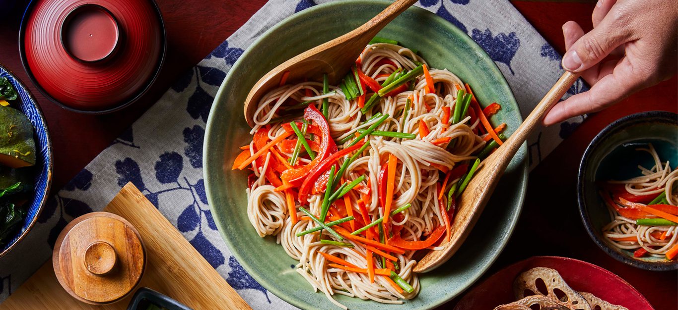 Soba Noodle Salad in a light blue bowl being tossed by a wooden fork and spoon