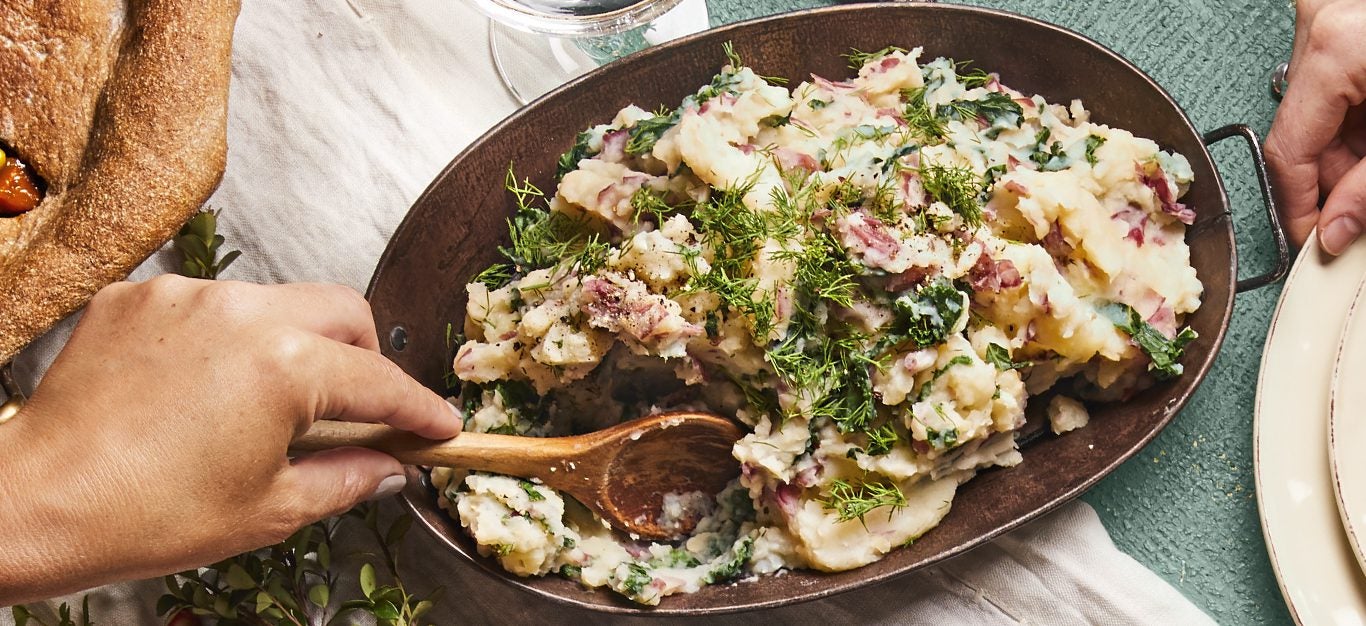 Mashed Potatoes with Kale and fresh dill in a metal serving dish being stirred by a woman's hand