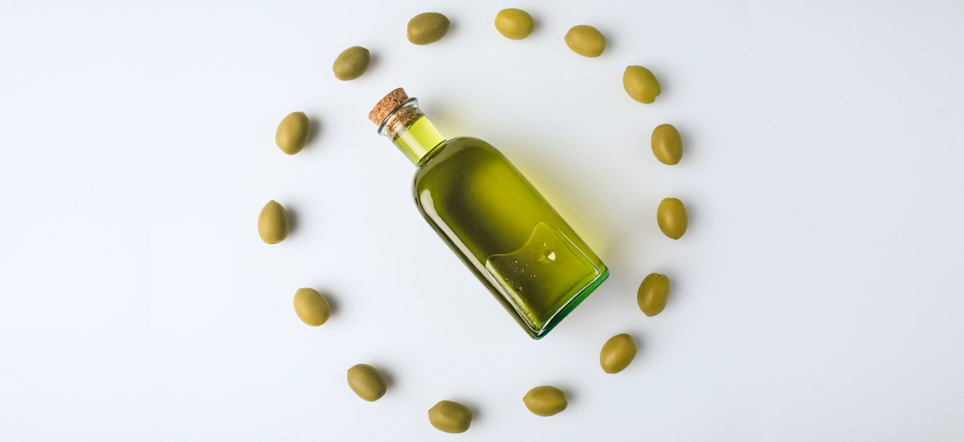 A corked clear glass bottle of olive oil lies on its side surrounded by a circle of green olives