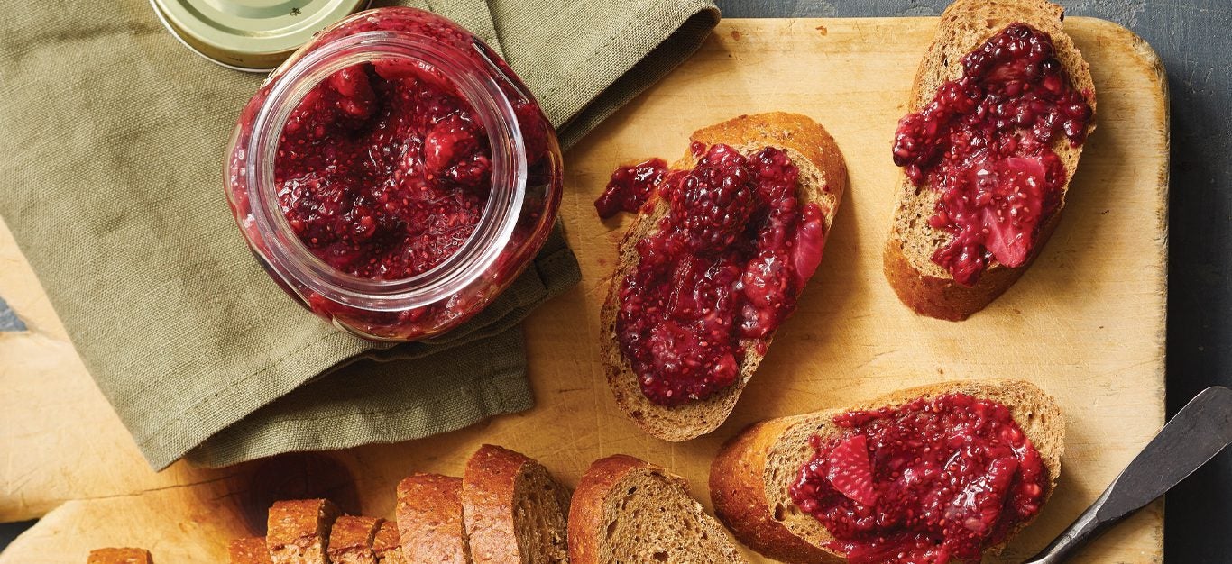 Homemade mixed berry jam spread on a few slices of whole grain bread with a jar of it off to the side