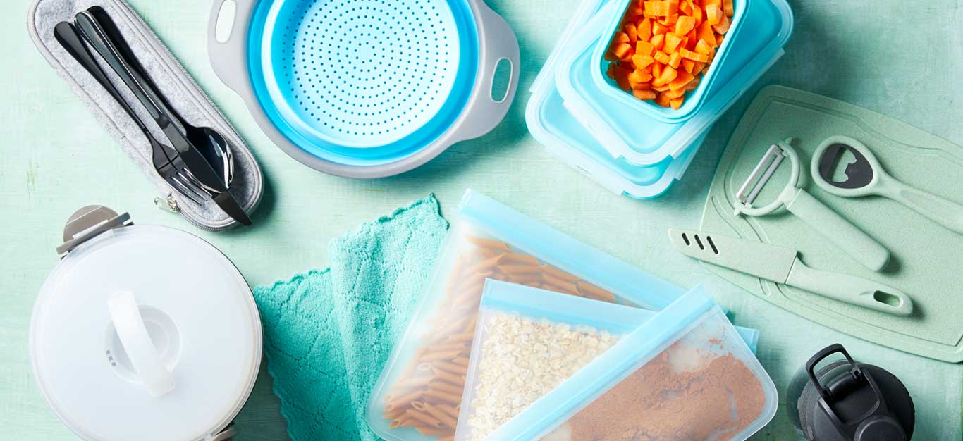 Utensils, resusable bag filled with grains and pasta, a colander, and glass tupperware filled with carrots on light blue tabletop