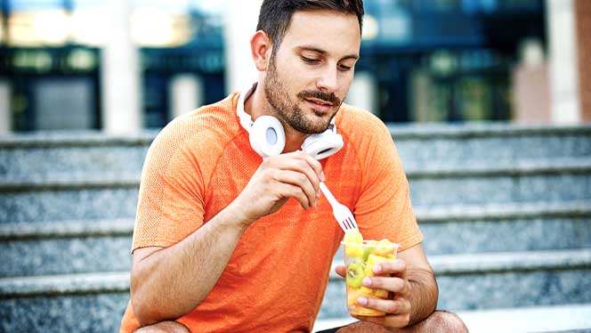 Man in orange shirt sits on steps eating a cup of fruit with headphones around his neck