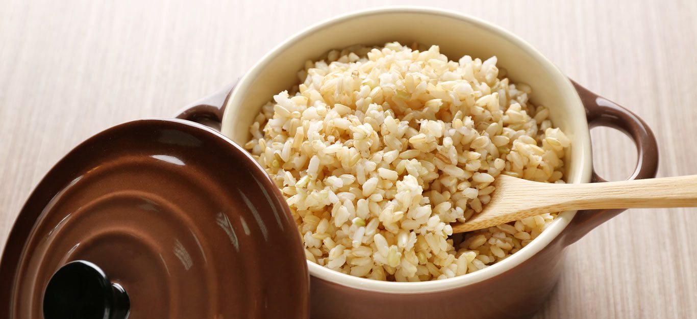 Brown ceramic saucepan full of cooked brown rice, with a wooden spoon nestled beneath the rice