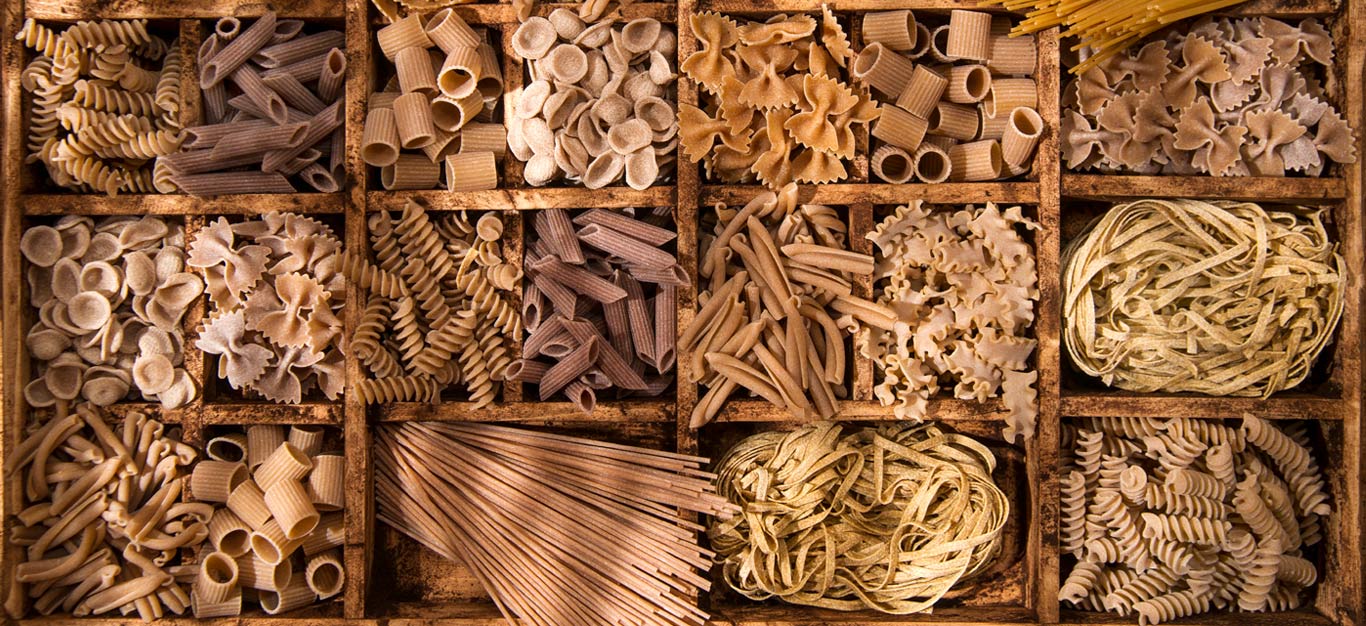 An array of whole wheat pasta shapes in a wooden crate, with compartments dividing each shape, including penne, orecchiette, spaghetti, and rotini