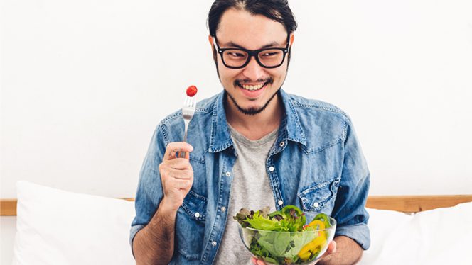 Young man wearing glasses holds a fork up with one hand and a bowl of green salad in the other, smiling