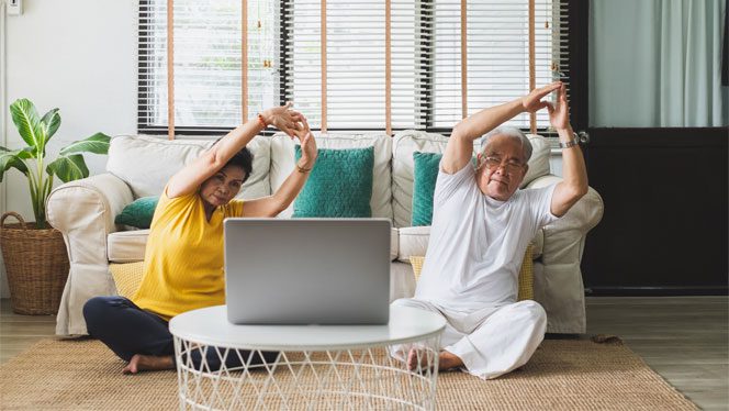 Two older Asian people sitting on the floor in front of a laptop stretching to an online qigong video