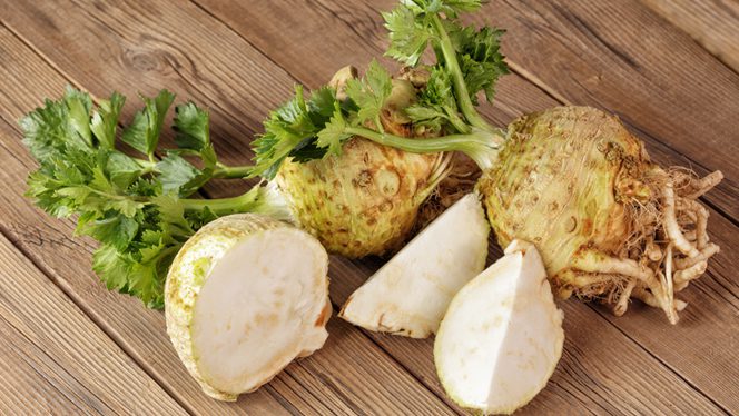 Two knobs of celeriac (celery root) on a wooden surface, with one knob cut into wedges showing the white flesh inside, and the other knob whole, with visible roots coming out of the bottom