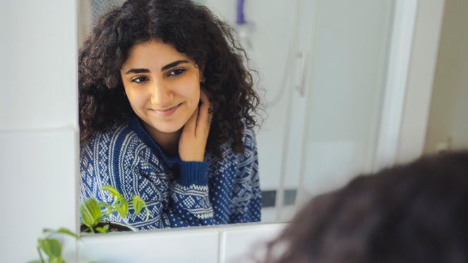 A young woman with curly hair looks into the bathroom mirror and smiles