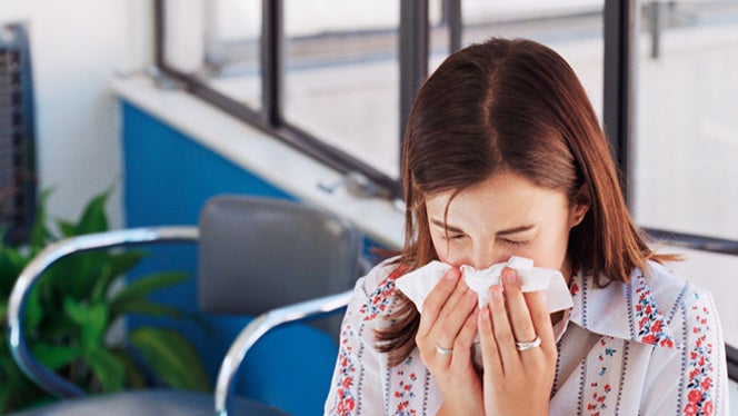 Woman sneezing standing inside an office near an open window from seasonal allergies