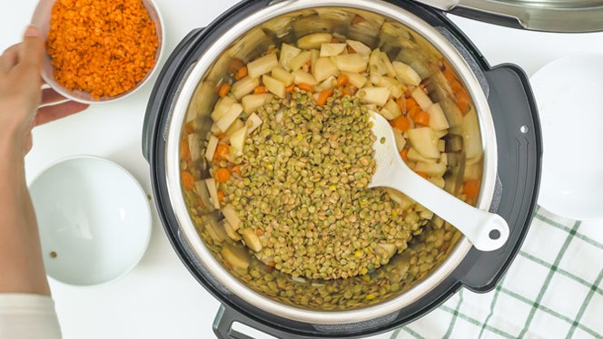 An overhead shot into an Instant Pot or multicooker with red and green lentils and potatoes in it, and a hand holding a bowl of shredded carrots about to empty into an Instant Pot