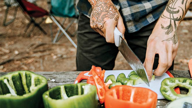 Man with tattoos on forearms slices bell peppers on a cutting board at a campsite