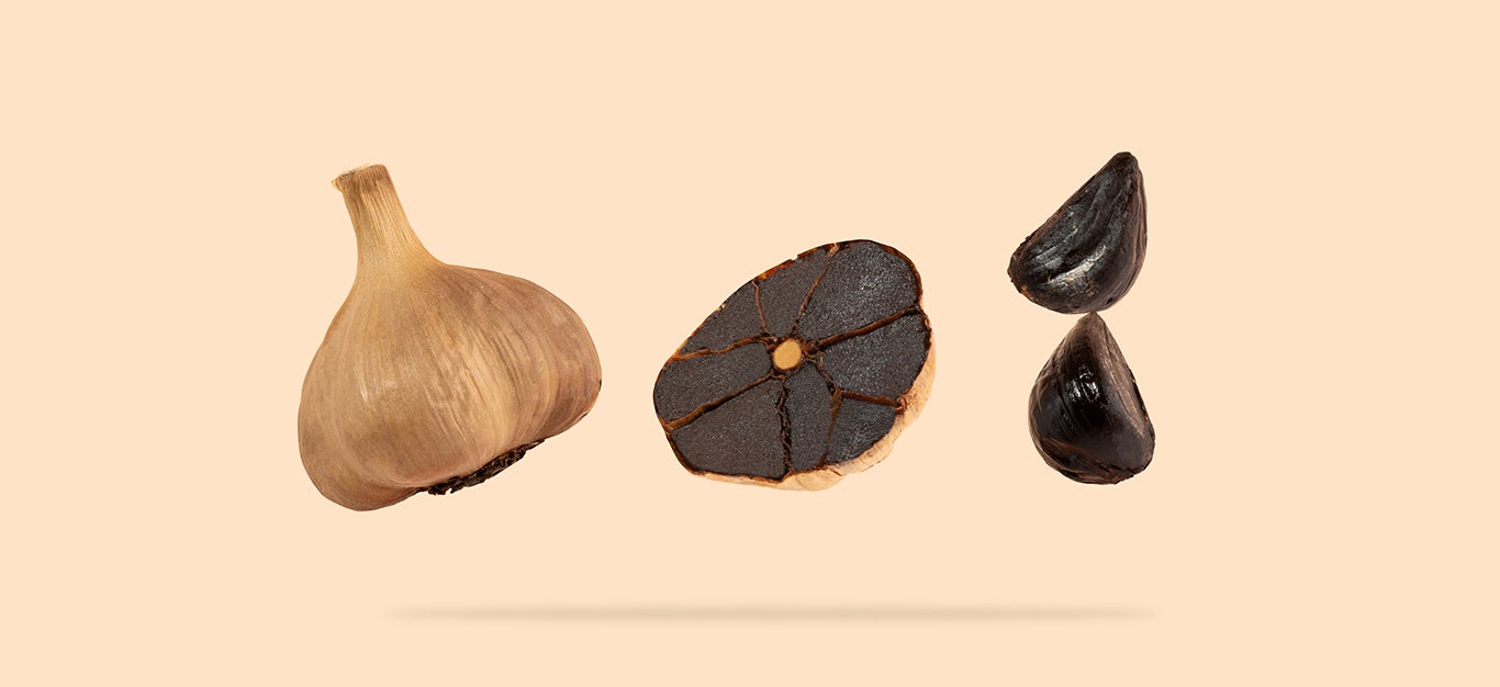 A whole head of black garlic shown next to a sliced head of black garlic and two peeled cloves black garlic, isolated on a light peach background