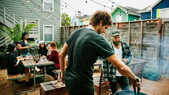 Two men in their 30s stand around a smoking grill cooking meat in a backyard, while in the background a group of friends sits around a picnic table drinking