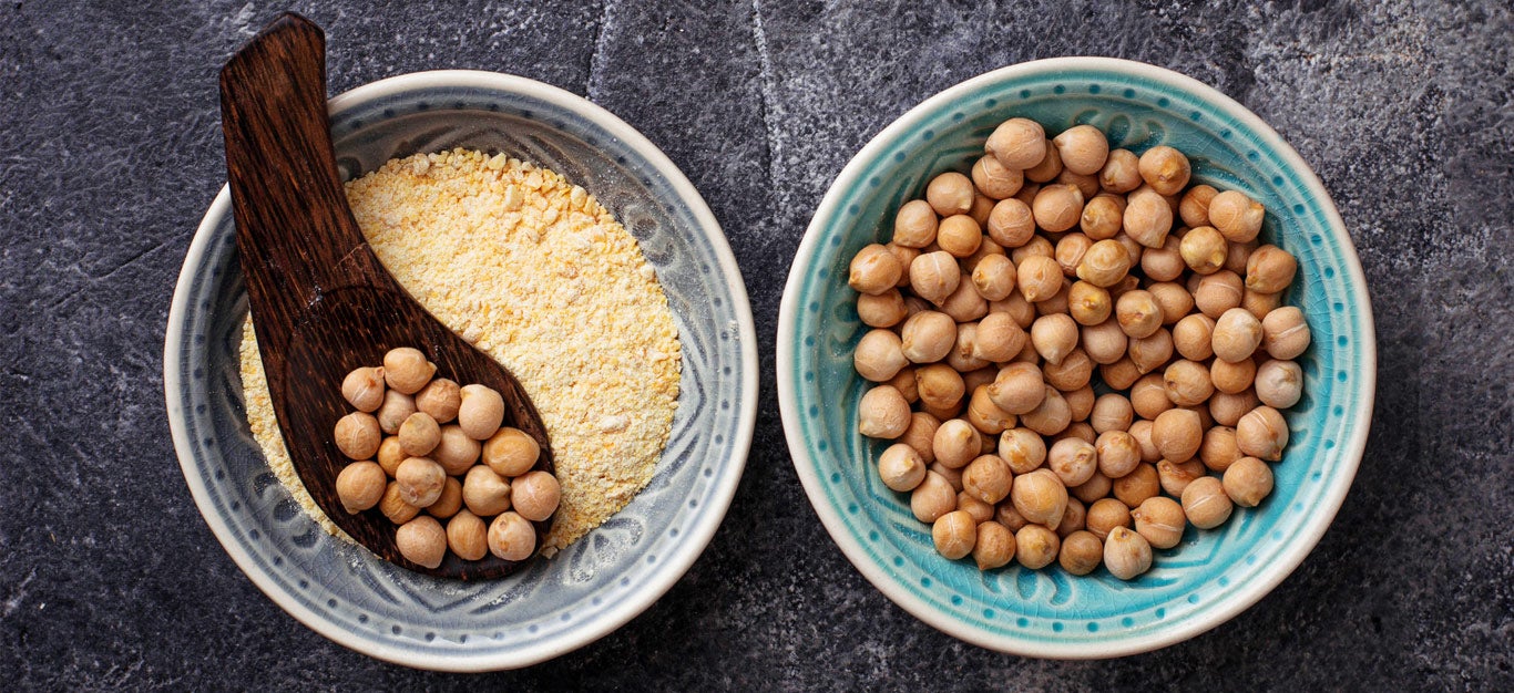 Photo of bowl of ground chickpea flour beside a bowl of whole dried chickpeas, with a spoon full of dried chickpeas resting atop the bowl of chickpea flour